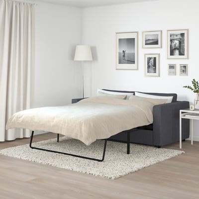 Modern bedroom with FINNALA sofa-bed, white bedding, black frame. White walls, curtains, wood floor. Black & white art. Spacious.