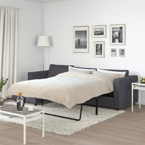FINNALA sofa-bed in dark grey transforms into a bed with black metal legs. Surrounded by photos, a floor lamp, and a side table on a patterned rug.