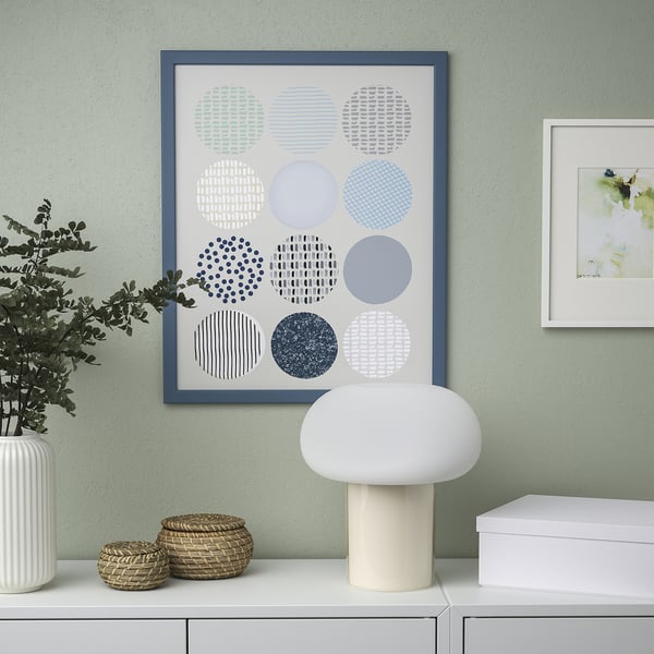 Modern gallery wall with blue-framed abstract art, white lamp, plant, and baskets.