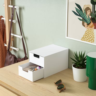 White mini storage chest with one drawer open, holding small items. Placed on a wooden surface near a coat rack and a framed poster.