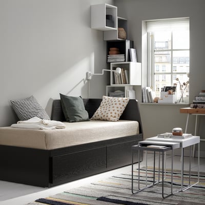 Modern living room with a black and beige FLEKKE sofa-bed, colourful rug, white nesting tables, and sleek wall shelves filled with books and decor.
