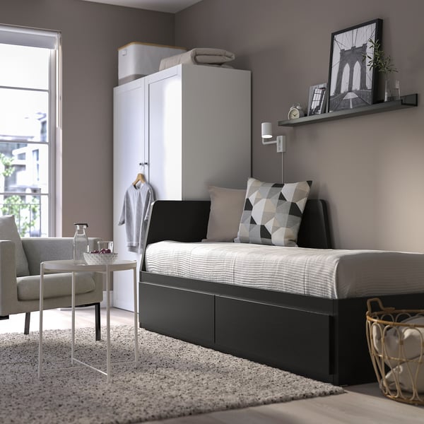 Modern bedroom with FLEKKE day-bed, storage drawers, tall cabinet, wall decor, armchair, and city view.