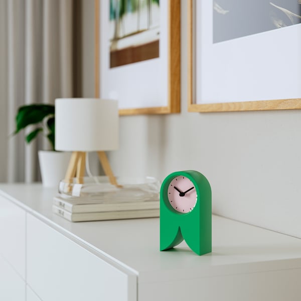 Small green FLODKRÄFTA clock sits on desk, silent, unique. Near lamp, books, plant, art. Lightweight, plastic.