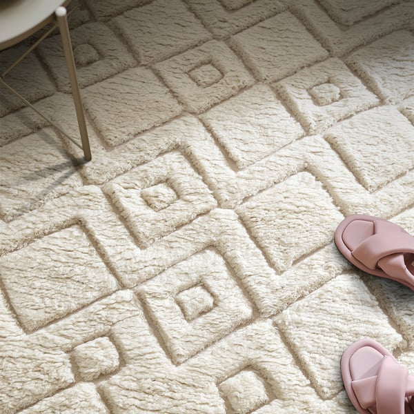 Beige textured rug with geometric pattern, soft to walk on, dries quickly, blends with furniture. Pink slippers to side.