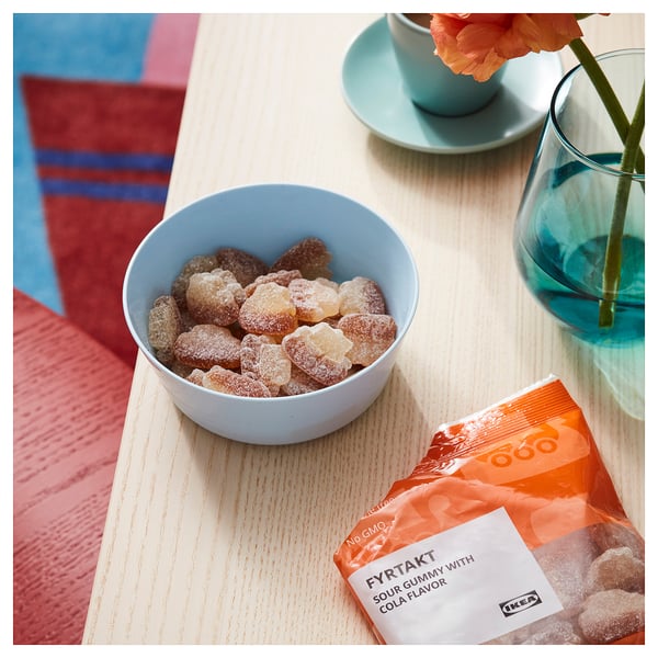 Blue bowl with candy, IKEA FYRTAKT cola gummies on wooden table, orange flower, teacup.