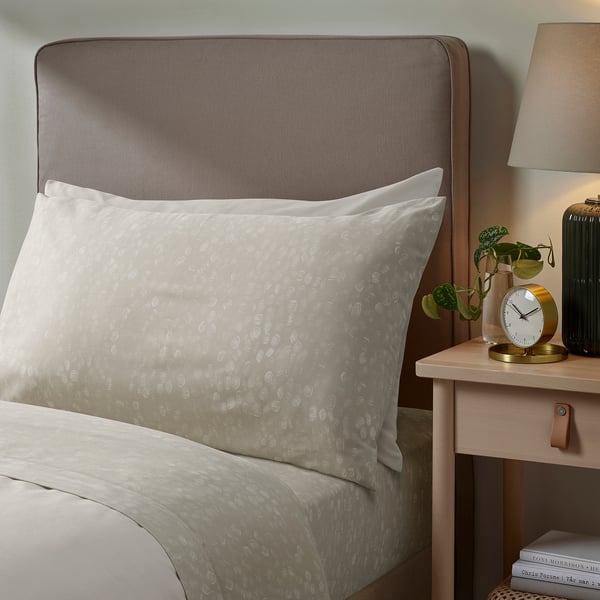 Bed with GINSTFÄLTMAL sheets, brown headboard, bedside table with clock, greenery, and lamp.