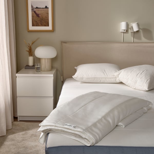 Minimalist bedroom with beige GRÄSROTSFLY duvet on large bed, white pillows, mushroom lamp, dried flowers, and framed poster.