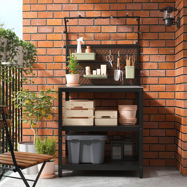 Black GRILLSKÄR outdoor storage against brick wall, holding garden tools and potted plants, larger than nearby chair.