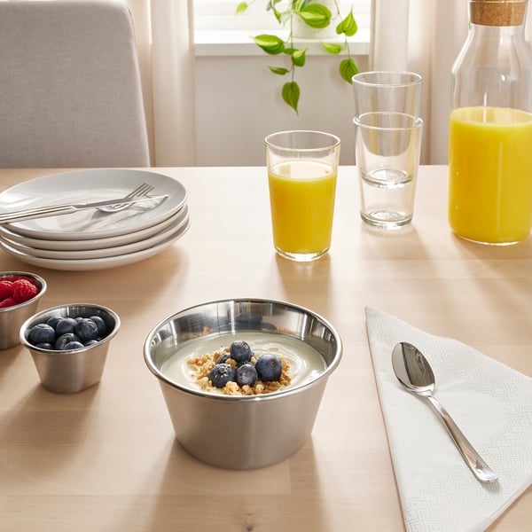 Table set with stainless steel bowl of yoghurt, berries, and granola. Glasses of orange juice and water, plates, cutlery, and a plant.