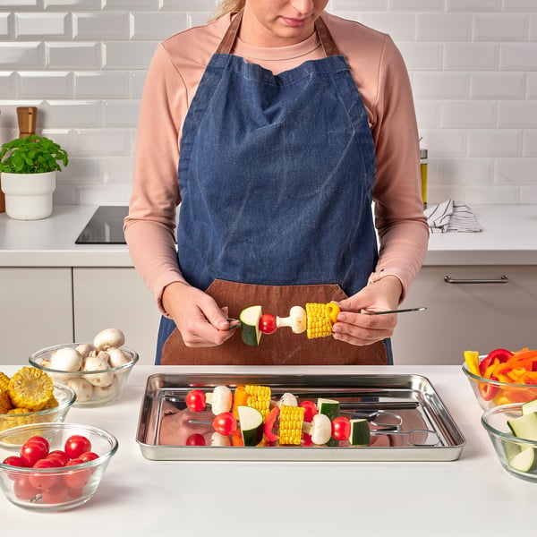 Person preparing skewers on metal tray with vegetables around.
