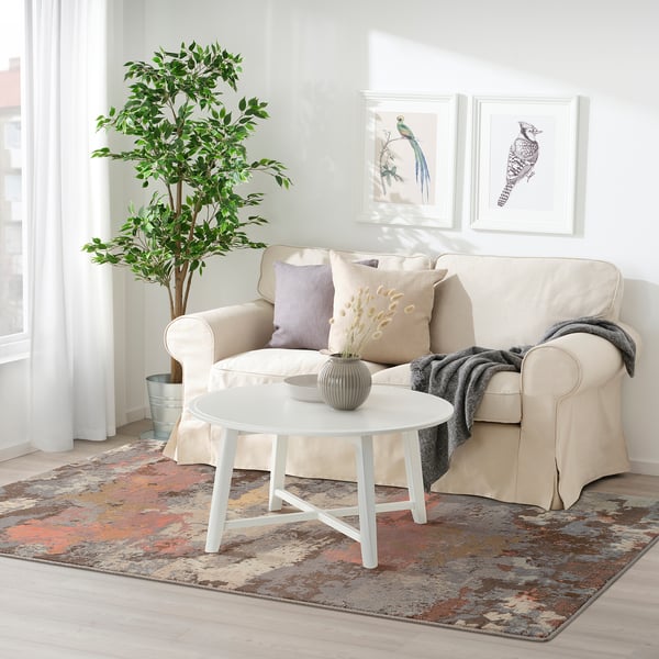 Modern living room with large beige sofa, two decorative pillows, grey blanket, white coffee table, multi-colored rug, potted plant, and wall art.