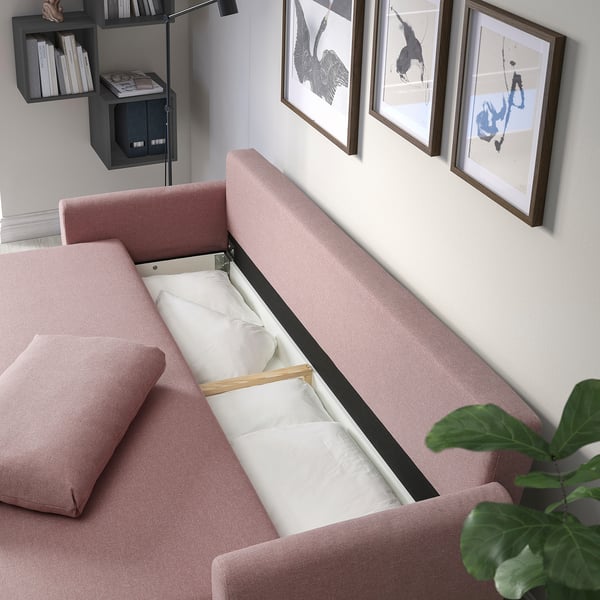 A pink sofa-bed is shown with white bedding inside, converting it into a full bed. Its against a wall with art and a shelf with books nearby.