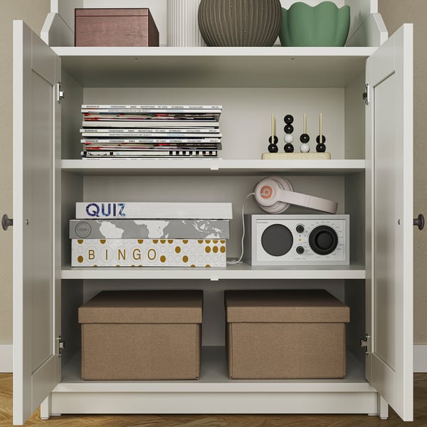White wooden cabinet with glass doors, holding books, games, headphones, and storage bins, neatly organised.