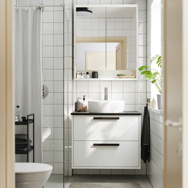 Modern bathroom with HAVBÄCK cabinet. Wall-mounted, minimalist design. Mirror, glass shower, sink with chrome tap. White tiled walls, grey towel.