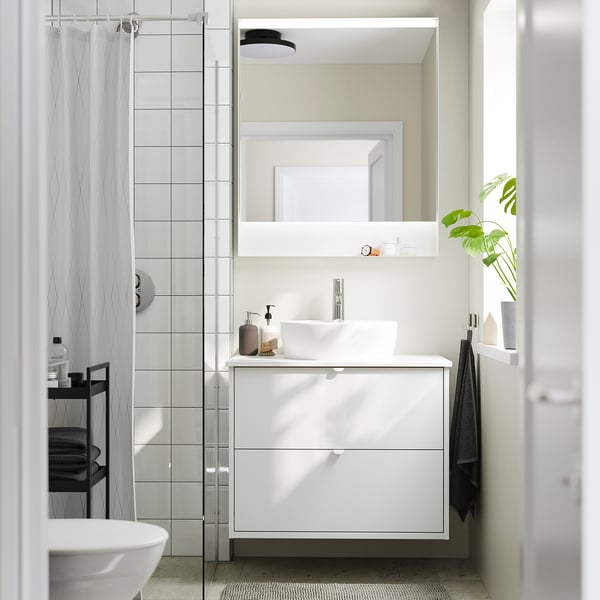 Modern white bathroom: HAVBÄCK unit, rectangular basin, mirror, glass shower, dark towels, and plant on shelf.