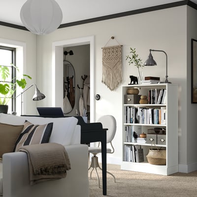 Bright living space with modern decor: white sofa, grey chair, wooden bookshelf with books, plants, and a black lamp.