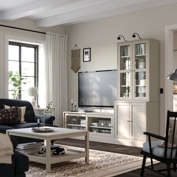 Modern living room with light brown wooden furniture including tv unit, cabinet, and chairs. Decorated with plants, cushions, and patterned rug.