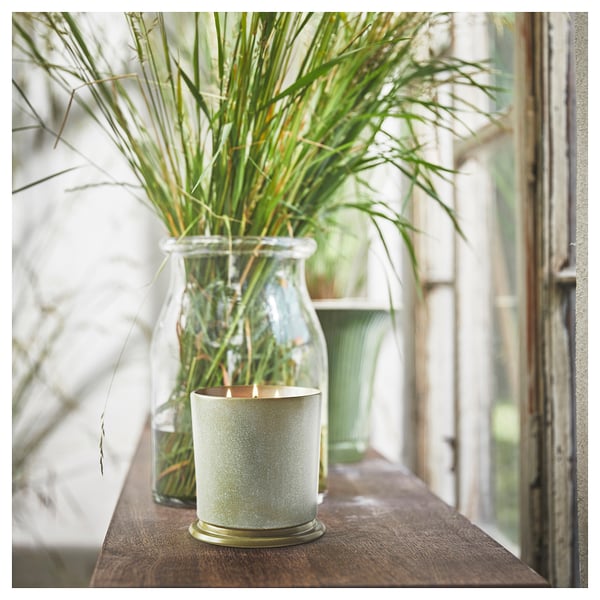 A lit candle in a textured white ceramic holder sits beside a glass jar filled with green plants on a wooden surface.