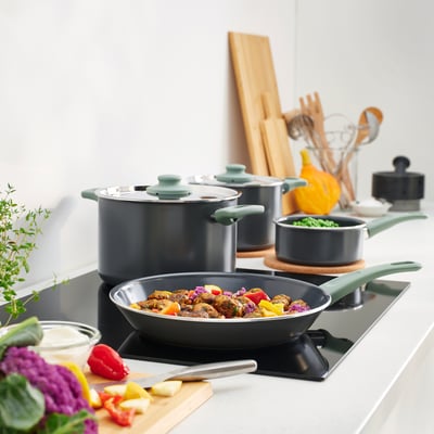 Modern kitchen with HEMLAGAD black cookware sets. Pots and pan with grey handles on induction stove. Vegetables and herbs nearby.