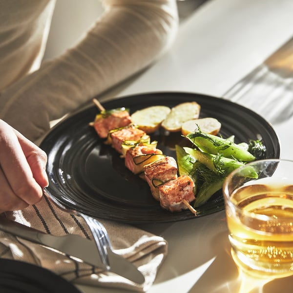 A hand reaches for a black plate with skewered food and vegetables, accompanied by a glass of beer.