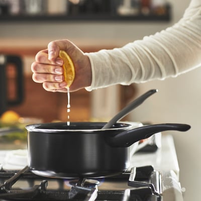 Image shows a person squeezing a lemon into a black saucepan on a stove. The saucepan has a non-stick coating and is placed over a lit burner.