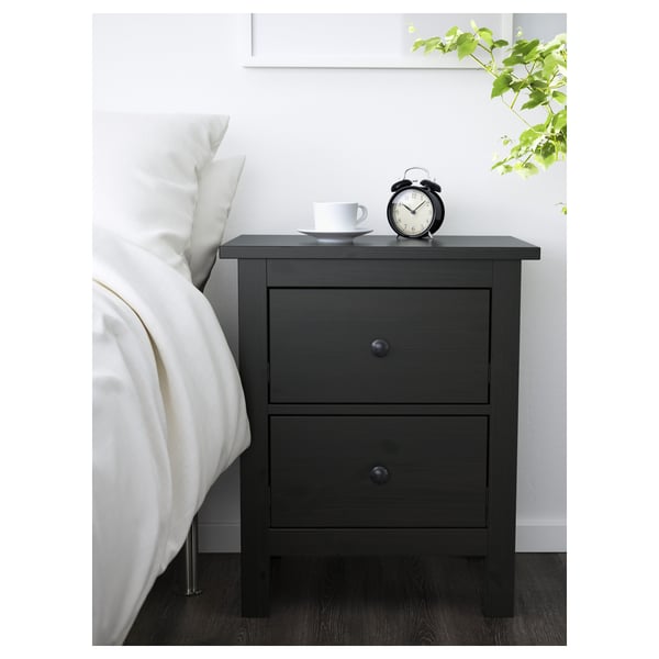 Bedroom setup with HEMNES bedside table. Dark wood, two drawers, smooth handles. On top, a clock and cup. Beside a white bed & green plant.