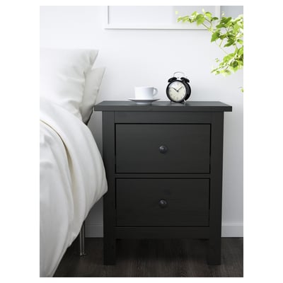 Bedroom setup with HEMNES bedside table. Dark wood, two drawers, smooth handles. On top, a clock and cup. Beside a white bed & green plant.
