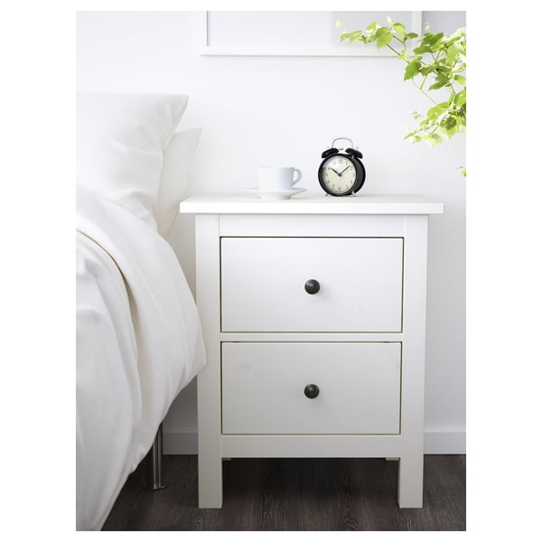 White HEMNES bedside table with 2 drawers beside bed, holding mug and alarm clock.