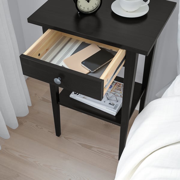 Black wooden bedside table with open drawer revealing a smartphone, notebook, and book. Clock and cup on top.