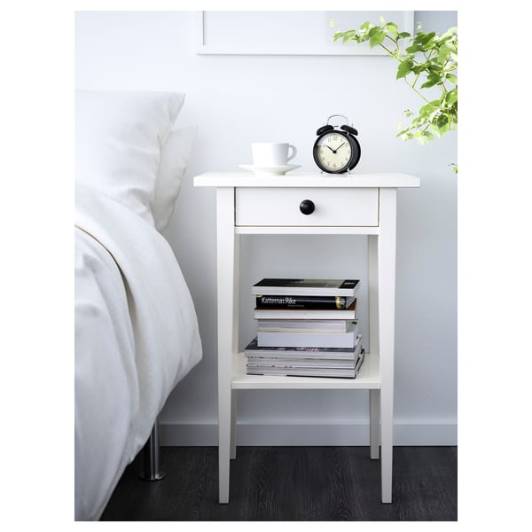 White HEMNES bedside table with black clock, coffee cup, books. Tall, slender, wooden design. Fits neatly beside large bed.