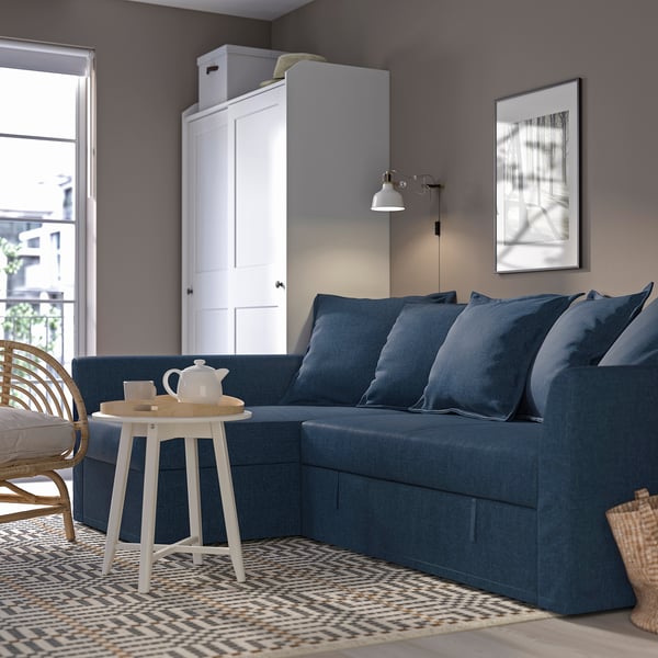 Modern living room with large blue HOLMSUND sofa, matching cushions, white side table, and wall lamp. Grey walls, patterned rug.