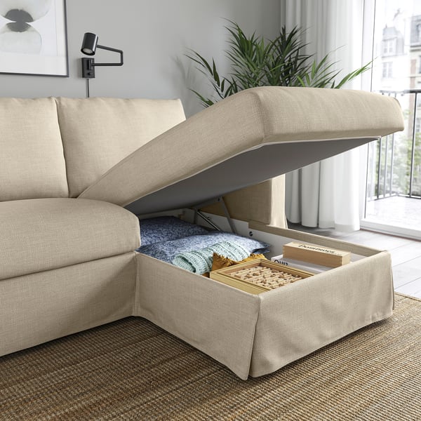 Neutral sofa with open storage beneath seat, revealing blanket and board games, set in bright room near large window.