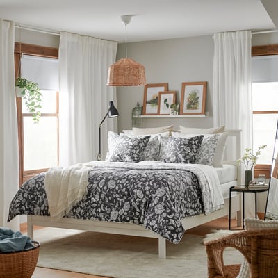 A black and white IDALINNEA bed set with floral comforter, white bed frame, and decorative elements in a bright, minimalist room.