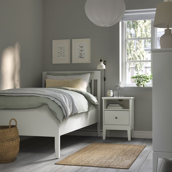 Minimalist bedroom with white bed, grey linens, yellow pillow. Art, bedside table, lamp, books, plant, rug, basket. Cosy and organised.