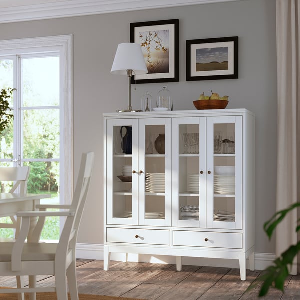 Glass-fronted white cabinet displays dishes, lamp and bowl of fruit on top. Nearby, wooden dining table with chairs against grey wall and framed art.