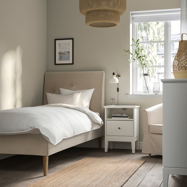 Beige IDANÄS bed with sloped headboard, white linen, bedside table, lamp, vase, large window, pendant lamp, cosy room.