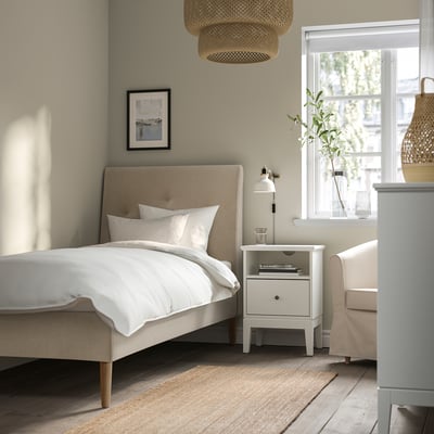 Beige IDANÄS bed with sloped headboard, white linen, bedside table, lamp, vase, large window, pendant lamp, cosy room.