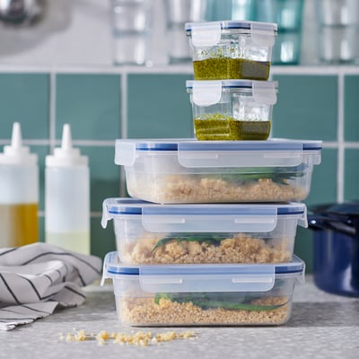 Stack of clear plastic food containers with white lids, holding grains and sauce, on a counter with kitchen items.
