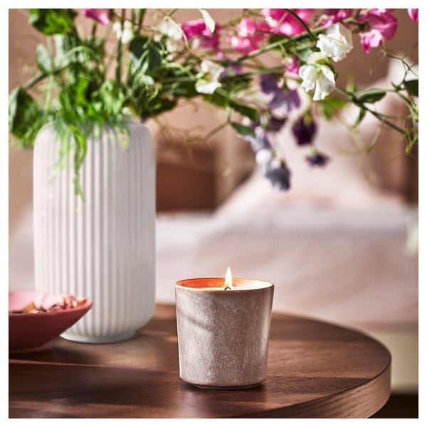 A lit grey ceramic candle on a wooden table with a white vase of colourful flowers beside it.