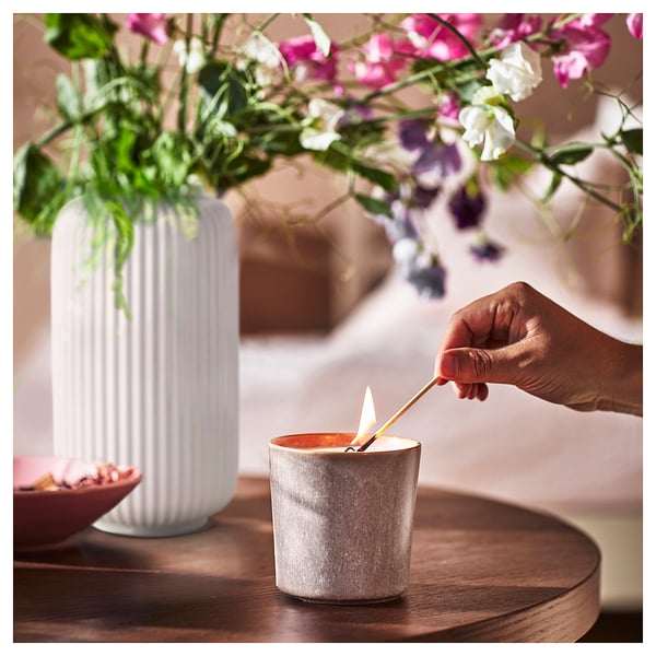 Lighting JÄMNMOD scented candle on wooden table, beside white fluted vase with colourful flowers.