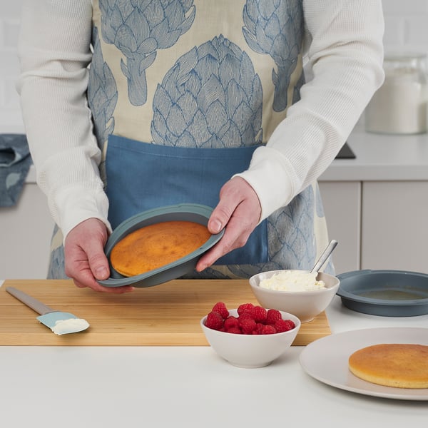 Person in apron holds cake pan, revealing baked cake. Counter has raspberries, icing, and another cake, depicting baking preparation.