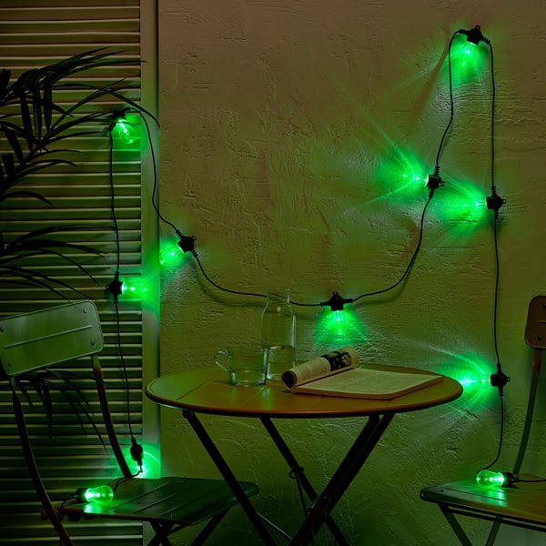 Green KASTTÅG lights hang, brightening room. Draped over table with glass, book, adding cosy festive ambiance.
