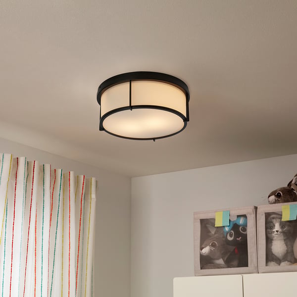 Black KATTARP lamp with frosted glass shade, above colourful curtains, cat photos, and toys on shelf.