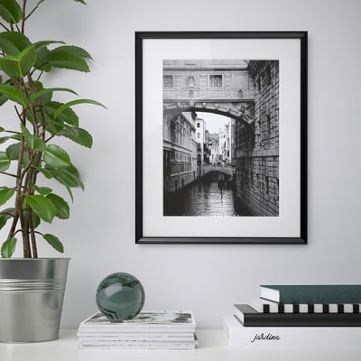 Black-framed a canal bridge on a wall. Next to a plant and stack of books.