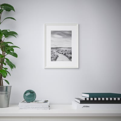 Framed path print on grey wall above shelf with magazines, turquoise sphere, and plant.