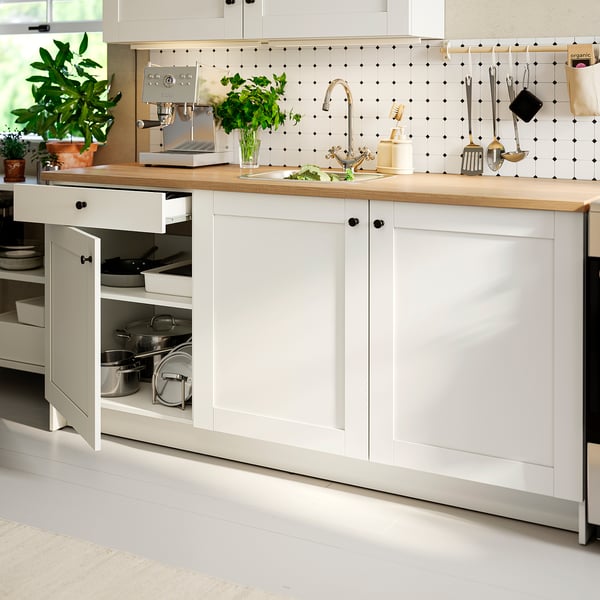 Modern kitchen with a white cabinet open, displaying pots inside, next to a countertop with a sink, tap, plant, and utensils.