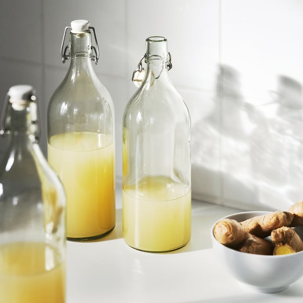 Three glass bottles with swing-top stoppers, filled with yellow liquid, on a white surface next to ginger roots.