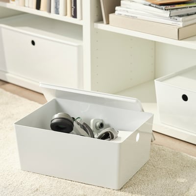 White storage box with lid on carpet, holding electronic devices, near bookshelf. Part of KUGGIS series.
