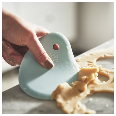Hand holds green LÄTTBAKAD spatula scraping light dough on white surface. Sleek, curved design in silicone and plastic.