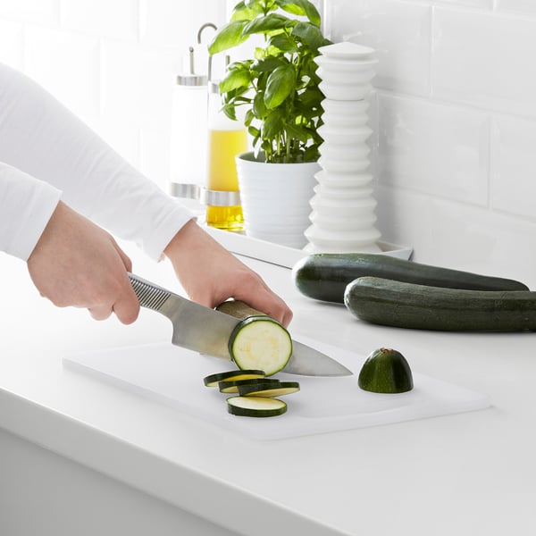 Person slices cucumbers on cutting board with knife, surrounded by plants and kitchen items.